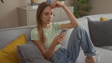 Yeşil gömlekli, kot pantolonlu, rahat bir oturma odasındaki kanepede oturan ve akıllı telefonunu düşünceli bir ifadeyle kullanan güzel bir kadın..