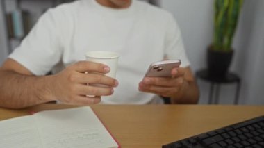 Ofiste elinde telefon ve kağıt bardakla bir adam var. Önünde bir defter ve bilgisayar var..