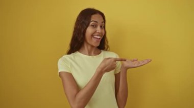 Genç bir kadın, Afro-Amerikalı, afallamış ve gülümsüyor, el ve işaret ediyor, izole edilmiş sarı arka planda tişört giyiyor.