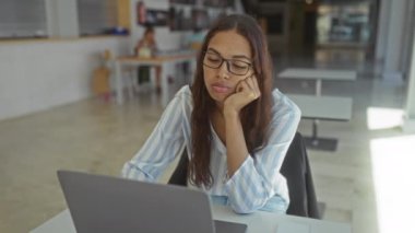 Kadın ofiste dizüstü bilgisayar başında çalışırken, gözlük ve çizgili gömlek giyerken, açık iç tasarımı olan modern bir iş yeri dekorunu sergilemekten yoruldu..