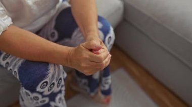 Evde oturan bir kadın, elleri kucağına bağlanmış, desenli pantolon giyiyor ve oturma odasında yüzük takıyor.