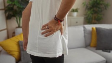 Modern bir oturma odasında sırtı ağrıyan orta yaşlı bir adam. Gri ve sarı yastıklar, kapalı bitkiler ve görünür rahat bir kanepe.