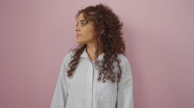 Esmer, kıvırcık saçlı, çizgili tişörtlü, soyutlanmış pembe bir arka plana bakan genç bir kadın.