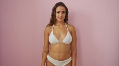 Bikinili genç İspanyol kadın, gülümsüyor ve pembe arka planda el işareti gösteriyor. Başarılı pozitif ifade ve izole ortamda kendine güven.