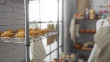 Fırında telefonla konuşan bir kadın. Rafta kekler ve arka planda beyaz tuğla bir duvar var.