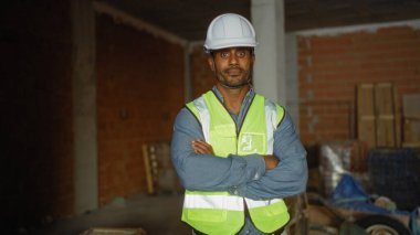 Handsome young man at a construction site wearing a hard hat and reflective vest stands confidently with arms crossed in an unfinished building clipart