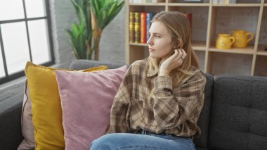 Evdeki kanepede oturan düşünceli sarışın kadın. Etrafı renkli yastıklarla ve kitaplıklarla çevrili..