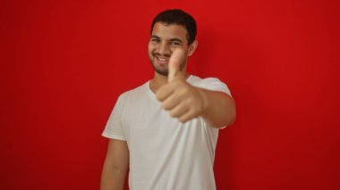 Yakışıklı İspanyol genç adam, izole edilmiş kırmızı arka planda dururken gülümsüyor ve başparmağını kaldırıyor.