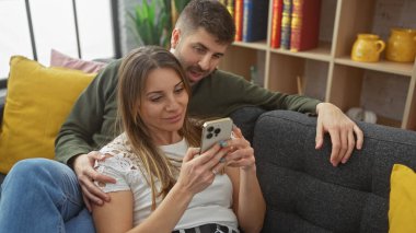 Bir çift, rahat bir oturma odasında dinleniyor, akıllı bir telefon tutan kadın, her ikisi de gülümseyerek, huzurlu bir ev hayatı öneriyor..