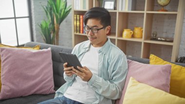 Gözlüklü genç bir Asyalı rahat ve modern oturma odasında tablet okumaya odaklanmış..