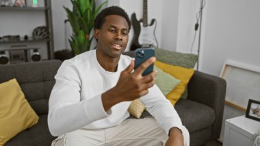 Modern oturma odasında akıllı telefon kullanan Afrikalı Amerikalı bir adam.