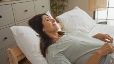 Olgun İspanyol kadın rahat bir yatak odasında huzur içinde dinleniyor, ev ortamında sakin bir atmosfer yaratıyor, dinleniyor, rahat giysiler giyiyor.