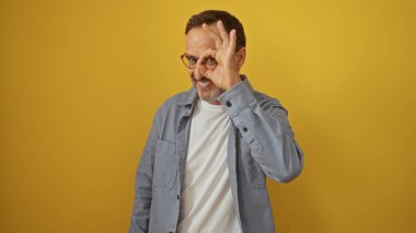 İspanyol adam, elini gözünün önünde, izole edilmiş sarı bir arka planın önünde gülümsüyor ve neşeli görünüyor..