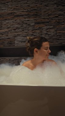 Rahat bir kadın modern bir banyoda taştan bir arka planı olan köpük banyosundan hoşlanır..