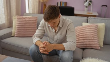 Oturma odasında oturan genç adam, başı eğik, evin iç dekorasyonunda rahat bir dekorla çevrili..