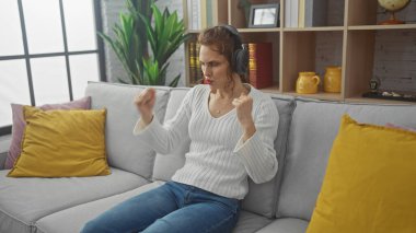 Düşünceli genç bir kadın kulaklıkla müzik dinliyor, renkli yastıklarla çevrili bir kanepede oturuyor..