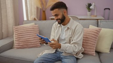 Hoş bir oturma odasında bej bir koltukta otururken tablet kullanan sakallı yakışıklı İspanyol bir adam.