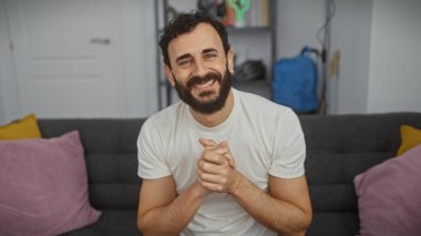 Modern bir oturma odasındaki kanepede oturan sakallı, beyaz tişörtlü, neşeli, orta yaşlı bir adam..