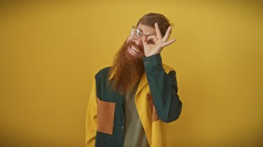 Gülümseyen sakallı genç adam sarı arka planda göze iyi bir jest yapıyor.