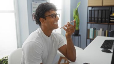 Ofiste telefonla konuşan genç adam kitaplıklar ve kapalı alandaki bitkilerle konuşuyor.