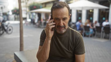 Şehir merkezinde orta yaşlı bir adam sokak kafesinde arka planda insanlarla telefonda konuşuyor.