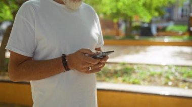 Güneşli şehir parkında beyaz tişört ve bilezik giyerken akıllı telefon kullanan bir adam.