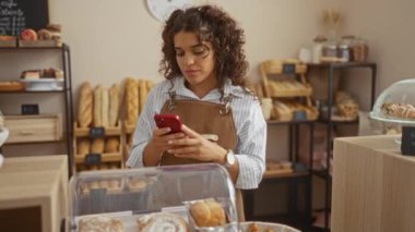 Fırında çalışan genç bir kadın akıllı telefon kullanıyor. Etrafı kruvasan ve ekmek gibi çeşitli fırınlarla çevrili. Raflarda sergileniyor.