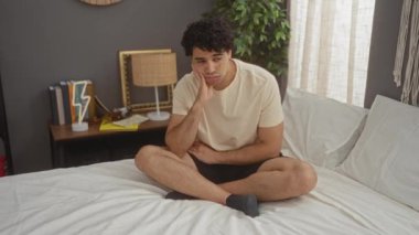 Genç İspanyol adam pijamayla yatakta oturuyor, gergin görünüyor ve yatak odasındaki stres ve stres yüzünden ellerini ısırıyor.