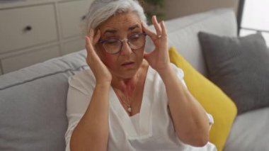 Oturma odasındaki kanepede oturan gri saçlı ve gözlüklü olgun bir kadın. Elleri şakaklarında stresli ya da acı içinde.