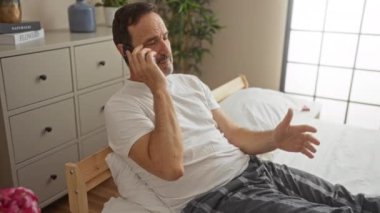 Orta yaşlı bir adam beyaz bir şifoniyerle rahat bir yatak odasında otururken telefonla konuşuyor.