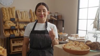 Fırında kamerayı işaret eden genç bir kadın ve eli ile buraya bir jest yapıyor, çeşitli fırınlanmış mallar ve ekmeklerle çevrili, kapalı yerlerde raflarda sergileniyor.