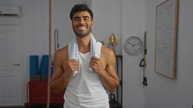 Arka planda ekipman bulunan bir spor salonunda boynunda havluyla güvenle gülümseyen genç bir adam.