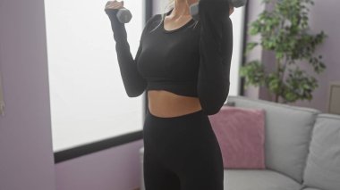 Genç bir kadın modern bir oturma odasında dambıllarıyla egzersiz yapıyor, spor yapıyor ve evde sağlıklı bir yaşam sürüyor.