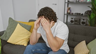 Stresli, kıvırcık saçlı, modern bir oturma odasındaki kanepede oturan, elleriyle yüzünü örten genç bir adam..