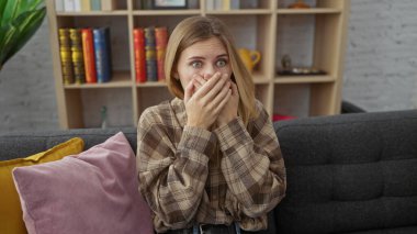Şaşırmış genç bir kadın, kitap dolu, rahat bir oturma odasında kanepede otururken elleriyle ağzını kapatıyor..
