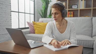 Kulaklıklı genç beyaz kadın dizüstü bilgisayar kullanıyor ve modern bir oturma odasında notlar alıyor.