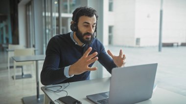 Kulaklık takan sakallı bir adam modern ofis ortamındaki bir video konferansında el kol hareketi yapıyor..