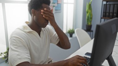 Ofisteki genç adam, bilgisayar masasında çalışırken eli yüzünde stresli görünüyor.