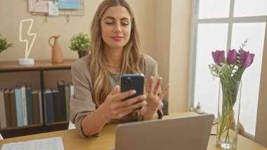 Odaklanmış bir kadın çiçek ve dcor ile ev ofis düzeninde dizüstü bilgisayarla akıllı telefon kullanır.