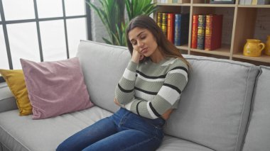 Düşünceli genç bir kadın kanepede oturur, rahat bir oturma odasında, dekoratif yastıklar ve kitaplıkla rahatlar..