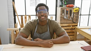 İyi donanımlı ahşap atölyesinde oturan, güvenlik gözlüğü takan gülümseyen Afrikalı Amerikalı adam..