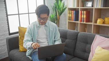 Asyalı adam rahat, modern bir oturma odasında dizüstü bilgisayar kullanıyor. Kitaplığı ve bitkileri var..