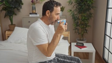 Orta yaşlı İspanyol bir adam rahat bir yatak odasında bir yatakta oturuyor, zarif dekore edilmiş bir iç mekanda bir kupadan yudumluyor ve bir ev ortamında bitki ve komodinle.