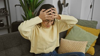 Gözlüklü stresli genç bir kadın koltukta oturur, elleri başının üstünde, endişe içinde ya da baş ağrısı içinde..