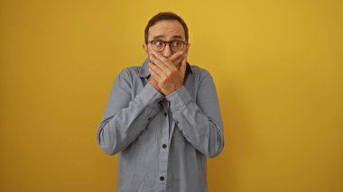 Orta yaşlı, ağzı iki eliyle kaplayan İspanyol bir adam, parlak sarı bir zemin karşısında gözlüklü ve mavi gömlekli, şaşırmış bir ifade sergiliyor.