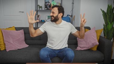 Günlük kıyafetli sakallı bir adam, kapalı bir kanepede oturmuş neşeli bir ruh hali içinde yedi parmağını gösteriyor..