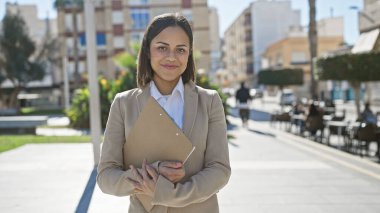 Elinde dosya tutan genç İspanyol iş kadını güneşli bir şehir sokağında güvenle duruyor..