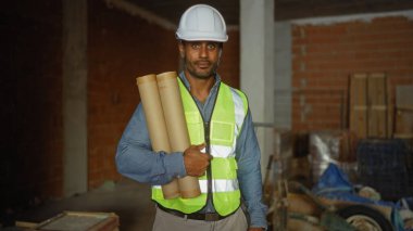 Şantiyedeki genç adam güvenlik yeleği ve başlık giyerken elinde planlar tutuyordu.