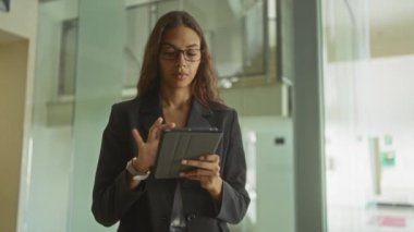 Ofiste gözlüklü ve spor ceketli bir kadın, modern bir iş yerinde çalışırken tablet kullanıyor.