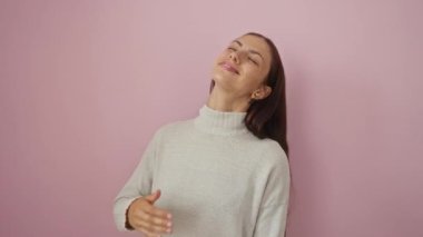 Açık renkli bir süveter giyerken pembe bir arka planda gülen ve gülümseyen genç bir kadın..